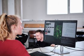 Eine blonde Frau sitzt an einem Bildschirm, auf dem die Perspektive des Fahrassistenten zu sehen ist. Diese zeigt das Gesicht eines Mannes, der in die Rückfahrkamera des Assistenten schaut