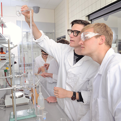 2 Studenten in Schutzbrillen und Kitteln schütten Flüssigkeit aus großer Höhe aus Reagenzglas in Behälter