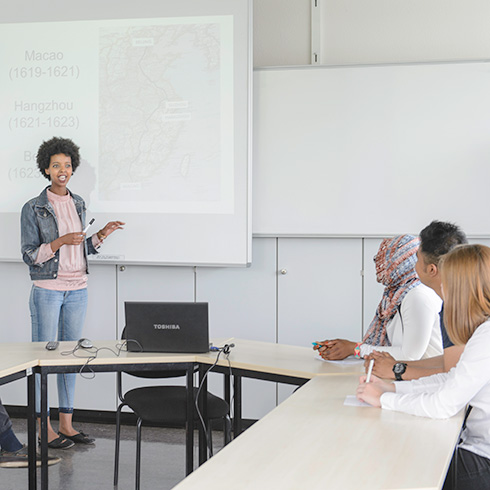 Eine Studierende steht vor einer Leinwand und hält einen Vortrag, drei weitere Studierende sitzen rechts am Tisch und schauen zu.