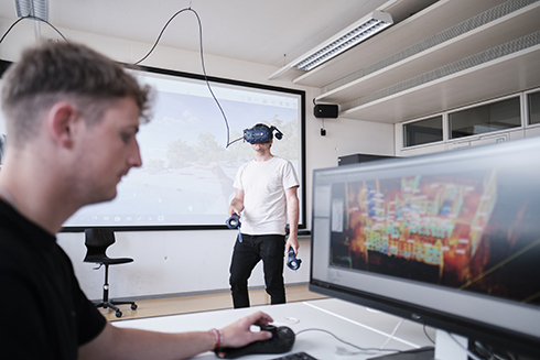 Student arbeitet am PC im BIM-Labor, Student mit VR-Brille im Hintergrund