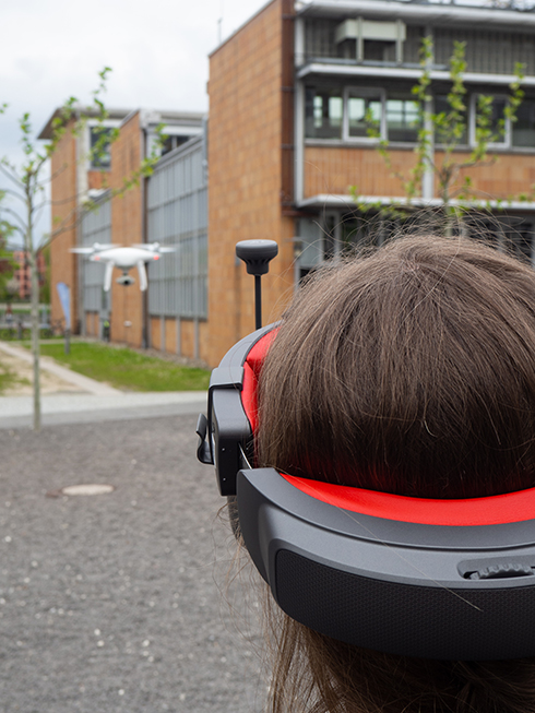 Studierende mit VR-Brille auf dem Campus, Flugdrohne fliegt im Hintergrund