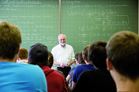 Ein Professor steht lächelnd vor den Studierenden, im Hintergrund ist die Tafel mit mathematischen Formeln zu sehen