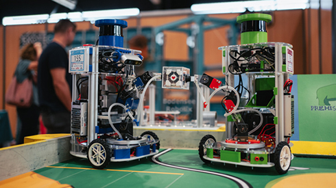 zwei Roboter auf einer Spielfläche, zwischen Ihnen ein Bauklotz mit Herz, im Hintergrund Personen