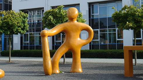 gelbe Figur mit Gewicht auf linkem Bein, rechtes Bein etwas abgesetzt angewinkelt aufgestellt, linker Hand in der Hüfte, rechte Hand hält einen Stab, der bis zum Moden reicht, rechts auf dem Bild ein Tischbein, Hintergrund ein Gebäude und Bäume, Boden Schotter 