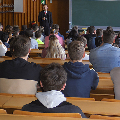 Viele junge Menschen sitzen in an langen Bankreihen in einem Raum und sind von hinten zu sehen. Vorn, neben einer Tafel, steht eine Person, die zu reden scheint.