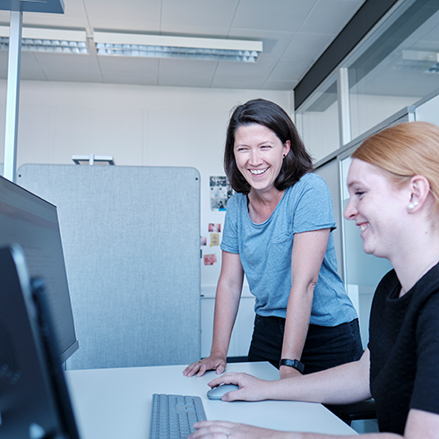 eine Person sitzt am PC, eine andere steht neben der Person am Tisch und schaut zu ihr herüber