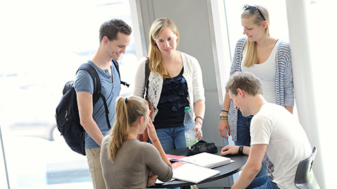 fünf Personen, zwei davon sitzen an einem Tisch, die anderen drei stehen um den Tisch, Unterlagen liegen auf dem Tisch