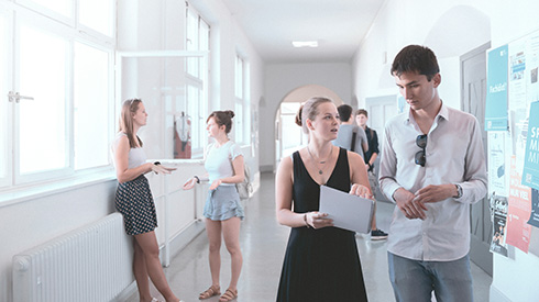 zwei junge Personen gehend und sich unterhaltend auf einem Flur, eine Person hat Unterlagen in der Hand, im Hintergrund zwei Personen die sich unterhalten, eine davon an die Wand vor einem offenen Fenster angelehnt