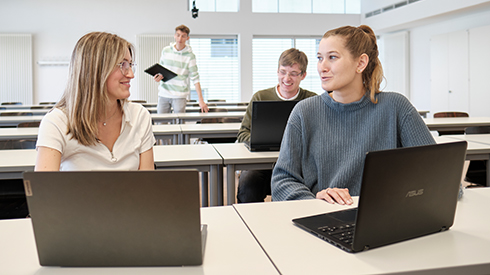 Zwei Personen sitzen an Tischen und sind sich zugewandt, vor ihnen Laptops. Im Hintergrund sitzt eine weitere Person am Laptop, dahinter scheint eine Person mit Laptop in der Hand gerade vom oder zum Tisch zu gehen. Im Hintergrund Tischreihen und rechts im Bild Fensterfläche.