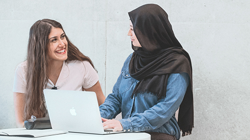 Zwei Personen stehen an einem Stehpult, auf dem ein Laptop steht. Sie unterhalten sich und die rechte Person hat die Hände auf der Tastatur.