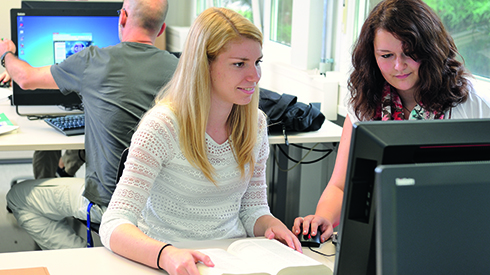 zwei Personen sitzen gemeinsam vor einem PC-Bildschirm, die Person links hat ein aufgeschlagenes Buch vor sich liegen, die andere die PC-Maus in der Hand. Beide schauen auf den Bildschirm. Im Hintergrund sitzt eine PC, die gerade den PC hoch- oder runterfährt.