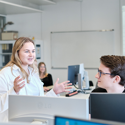 links steht eine Person, die anscheinend mit einer sitzenden Person spricht. Dabei gestikuliert die stehende Person. Im Vordergrund PCs von hinten, im Hintergrund PCs, eine Person und weiße Tafel.