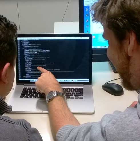 Zwei Personen von hinten vor einem Laptop mit Programmcode, eine Person zeigt auf eine Code-Stelle am Bildschirm