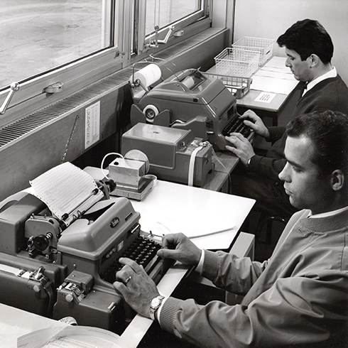 schwarz-weiß Foto, zwei Männer sitzen in einem Raum vor einem Fenster mit schreibmaschinenartigen Geräten