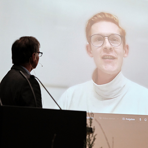 links ist eine Person von seitlich hinten vor einem Rednerpult zu sehen, rechts auf einer Leinwand eine weitere Person