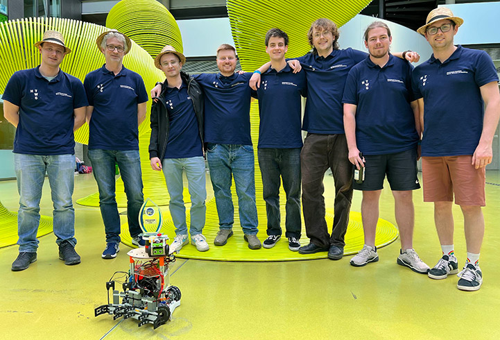 Acht Personen mit dunkelblauem Polo und Strohhut stehen nebeneinander, vor ihnen steht ein Roboter.