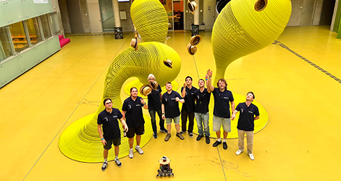 Acht Personen mit dunkelblauen Shirts werfen einen Strohhut in die Höhe. Vor ihnen steht ein mobiler Roboter. Im Hintergrund sind große gelbe astrakte Figuren auf einem gelben Boden platziert.