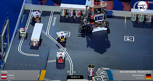 Robotik-Spielfläche mit dem HTWG-Roboter und kleineren Robotern, links unten ist TDA, Wels, Austria mit Flagge eingeblendet, rechts unten Bodensee RobotiX, Konstanz, Germany mit Flagge