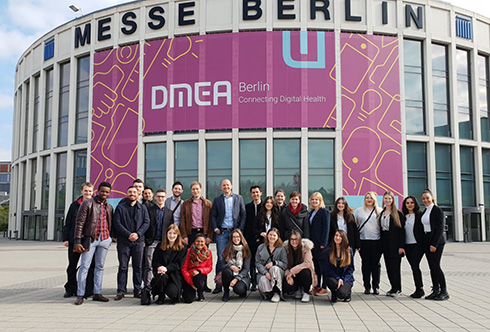 größere Personengruppe steht und kniet vor einem halbrunden Gebäude auf dem "Messe Berlin" und "DMEA" zu lesen ist