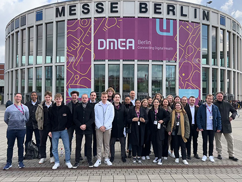 Eine Gruppe mit vielen Personen steht vor einem Gebäude, auf dem "Messe Berlin" steht und auf pinkem Hintergrund "DMEA"