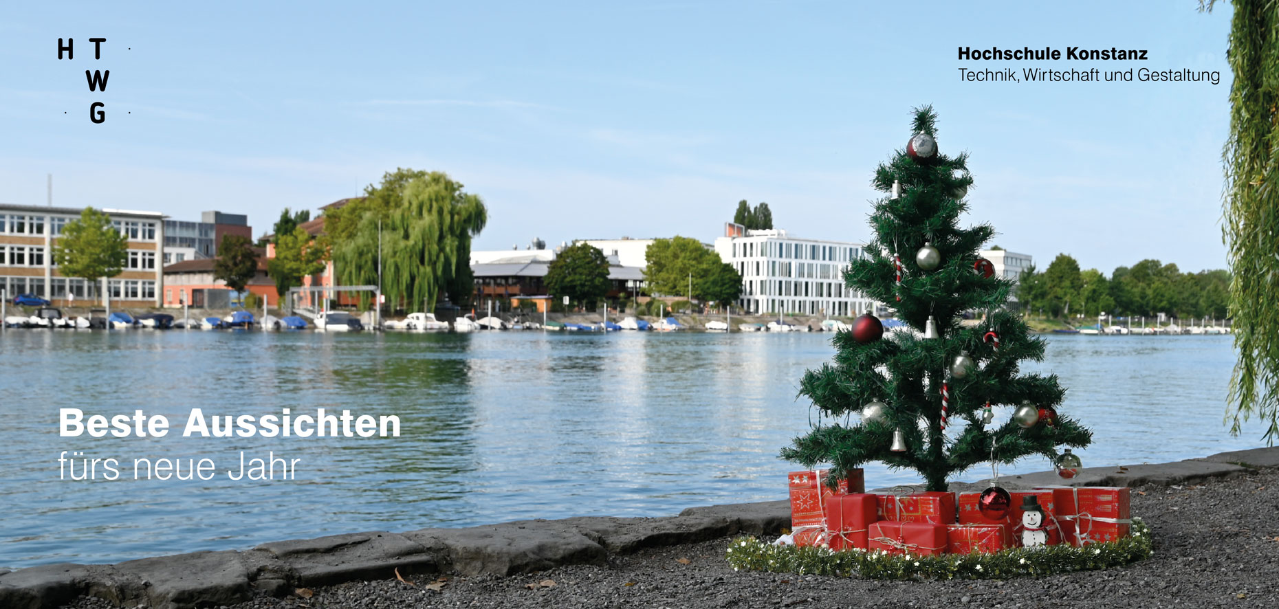 Geschmückter Weihnachtsbaum bei sommerlichem Wetter am Ufer, darunter etliche Pakete; auf der anderen Rheinseite sieht man HTWG-Gebäude; Text: Beste Aussichten für das neue Jahr sowie HTWG-Logo und vollständiger Hochschulname