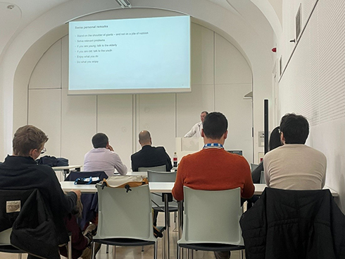 Vortragsraum, Projektionsfläche ist zu sehen sowie eine sprechende Person und die Rückenansicht mehrerer sitzender Teilnehmende