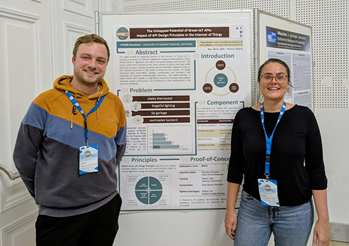 Zwei Personen seitlich neben einem personenhohen Aufsteller an dem ein Poster hängt.