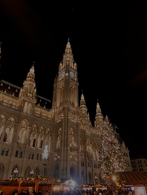 ein altes beleuchtetes großes Gebäude in der Dunkelheit, davor ein beleuchteter Tannenbaum und Marktstände