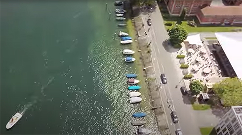 linke Bildhälfte Wasser, ein Boot fährt unten links aus dem Bild; Bildmitte: viele Boote haben am Ufer angelegt, dahinter Bürgersteig und Straße, dann verschiedene angeschnittene Gebäude, vor einem sind viele Sonnenschirme auf Sandstrand platziert 