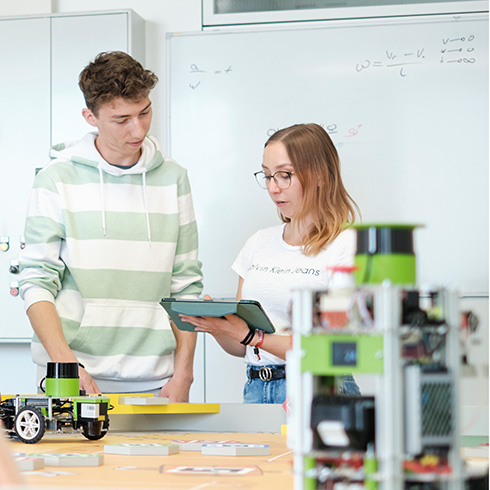 Rechts vorne im Bild ist unscharf ein Roboter auf einer ockerfarbenen Eurobot-Spielfläche zu sehen. Hinter der Spielfläche stehen zwei Personen, die beide auf ein Tablet blicken, das die Person rechts im Bild in der Hand hält.