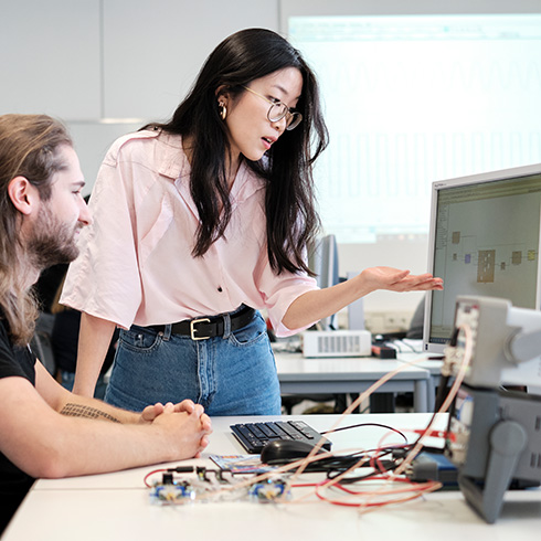 Auf einem Tisch stehen rechts technische Geräte mit Verkabelung, dahinter ein Bildschirm, auf dem was zu sehen ist. Eine Person sitzt vor dem PC, eine andere steht seitlich neben dieser und weist mit der Hand auf den Bildschirm, auf den beide blicken.