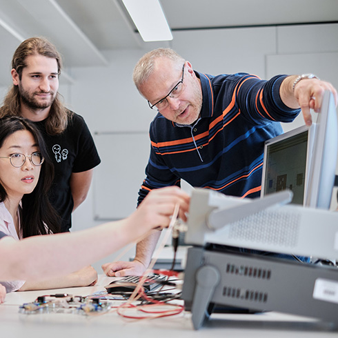 Im Vordergrund ist ein technisches Gerät mit Kabeln zu sehen, dahinter ein Bildschirm und eine Person, die mit dem Arm etwas auf dem Bildschirm anzeigt. Neben der Person schaut eine Person stehend und eine Person sitzend auf den Bildschirm und das Gerät.