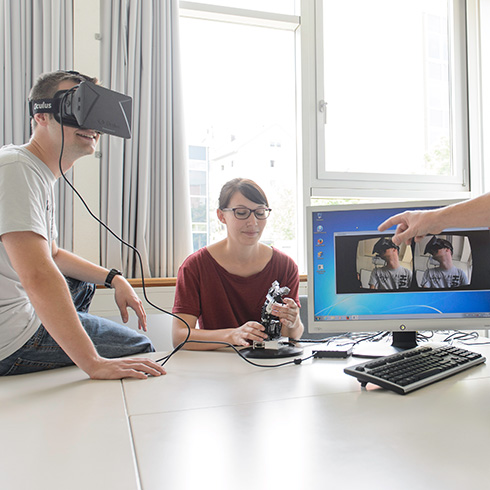 eine Person mit einer VR-Brille sitzt auf einem Tisch mit Blickrichtung nach rechts,rechts daneben sitzt eine Person auf einem Stuhl und hält etwas Technisches in der Hand, daneben steht ein PC-Bildschirm mit Tastatur, auf dem Bildschirm ist die Person mit VR-Brille zwei Mal zu sehen, Hand einer dritten Person zeigt von rechts auf das Bildschirmbild