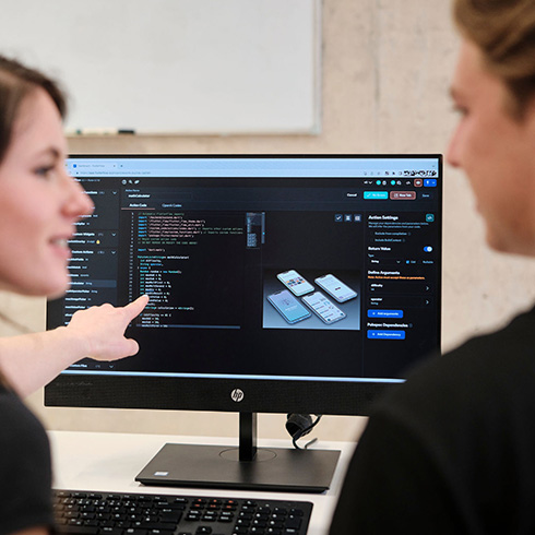 Zwei Personen sitzen vor einem Bildschirm. Beide Personen sind seitlich angeschnitten zu sehen. Die Person links zeigt auf den Bildschirm, während sie die andere Person anschaut. Auf dem Bildschirm sind Programmcode und Smartphone-Abbildungen zu sehen.