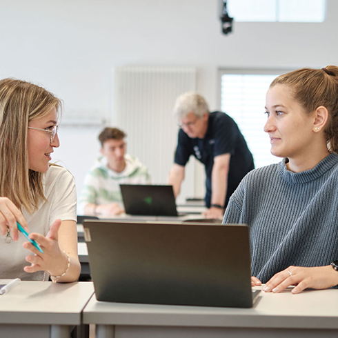 Zwei sich unterhaltende Personen (von vorn), vor ihnen ein Laptop. Im Hintergrund sind zwei weitere Personen an einem Laptop.