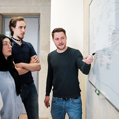 Drei Personen vor einem Whiteboard mit skizziertem Prozessablauf; eine Person zeigt auf ein Element.