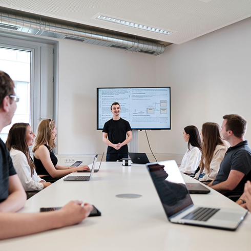 Sitzende Personengruppe am Besprechungstisch. Eine Person steht vorne neben einem Standbildschirm.