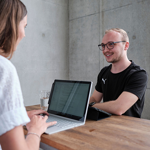 Zwei Personen die sich anschauen mit Laptop bzw. Tablet an einem Stehtisch aus Holz.