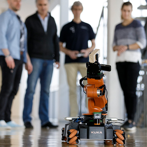 Vier Personen stehen nebeneinander, im Vordergrund ist ein schwarz-orange-farbener Roboter mit Greifarm auf dem Boden zu sehen. Der Greifarm umfasst einen hölzernen Klotz.