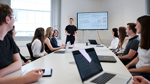 Eine Personengruppe sitzt an einem länglichen Tisch. Auf dem Tisch stehen einige Laptops und Tablets. Am Ende des Tischs steht eine Person und scheint zu den anderen zu sprechen. Neben der Person ist ein Standbildschirm.