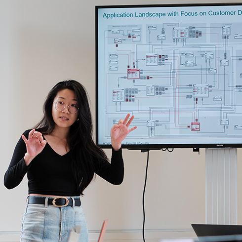 Studierende-Person-stellt-Applikationslandschaft-vor-Wirtschaftsinformatik-Studium-HTWG-Konstanz Eine Person steht neben einem Standmonitor, auf dem "Application Landscape with Focus on Customer Data" oberhalb einer sehr aufwändigen Abbildung steht. Die Person hat beide Unterarme gehoben und macht Handbewegungen.