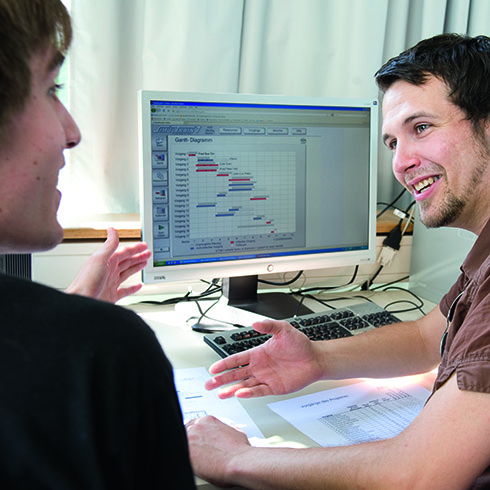 links ist angeschnitten eine Person zu sehen, die die linke Hand gestisch gehoben hat, rechts danaben eine Person, die diese anblick und den rechten Arm und Hand gestisch nutzt. Vor der Person liegt ein bedrucktes Blatt Papier, dahinter steht ein PC. Auf dem Monitor ist ein Diagramm abgebildet. 