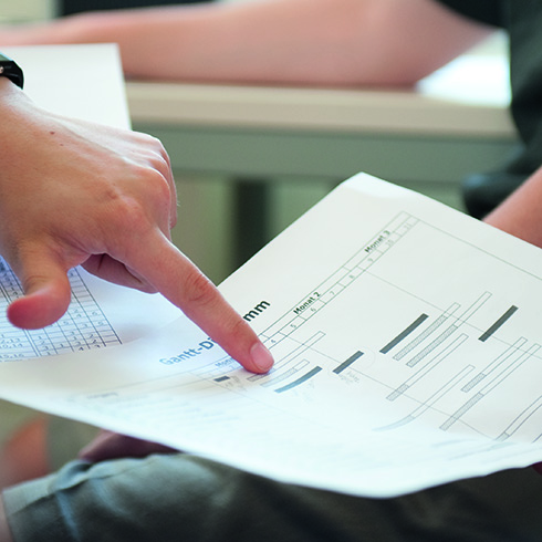 Links im Bild ist eine Hand zu sehen, deren Zeigefinger auf einen Balken im Gantt-Diagramm zeigt. Das Papier mit dem Diagramm scheint von einer anderen Person rechts gehalten zu werden, im Hintergrund sind die Arme einer weiteren Person zu sehen. 