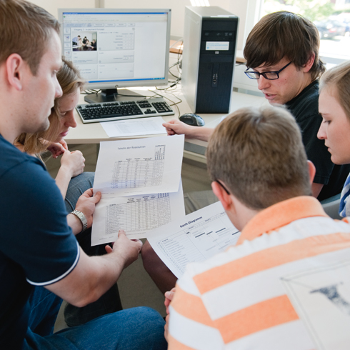 Drei junge Männer und zwei junge Frauen sitzen im Halbkreis vor einem Computer. Ihre Blicke sind auf zwei bedruckte Blatt Papier gerichtet, die zwei Personen aus der Gruppe in den Händen halten.. 