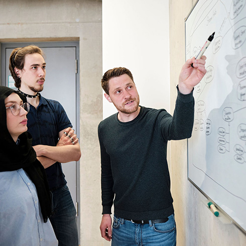 Drei Personen stehen vor einem Whiteboard, auf dem ein Prozessablauf aufgezeichnet ist. Eine Person zeigt mit einem Stift auf ein Element der Abbildung.