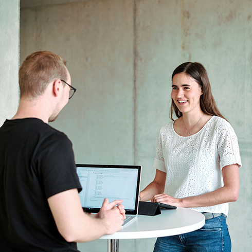 Zwei Personen stehen an einem kleinen runden Stehtisch. Die Person links ist von hinten zu sehen und hat einen Laptop vor sich stehen, die Person rechts ist von vorn zu sehen und hat ein Tablet vor sich liegen. Die beiden Personen schauen sich an und scheinen sich zu unterhalten.