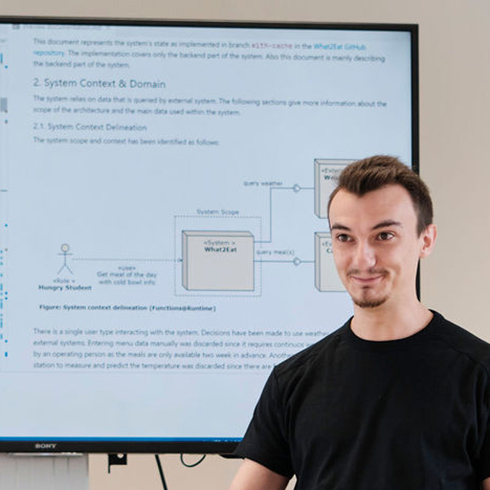 Eine Person steht vor einem Bildschirmhintergrund. Auf dem Bildschirm sieht man links Text und rechts eine schematische Darstellung einer Software-Architektur