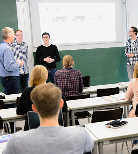 Studierende von hinten in einer Vorlesung. An der Tafel ein Dozent mit weiteren Studierenden im Gespräch