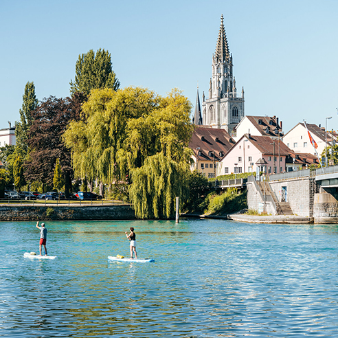 Two persons stand up paddeling on Lake Constance with the the cathedral of Konstanz in the background
