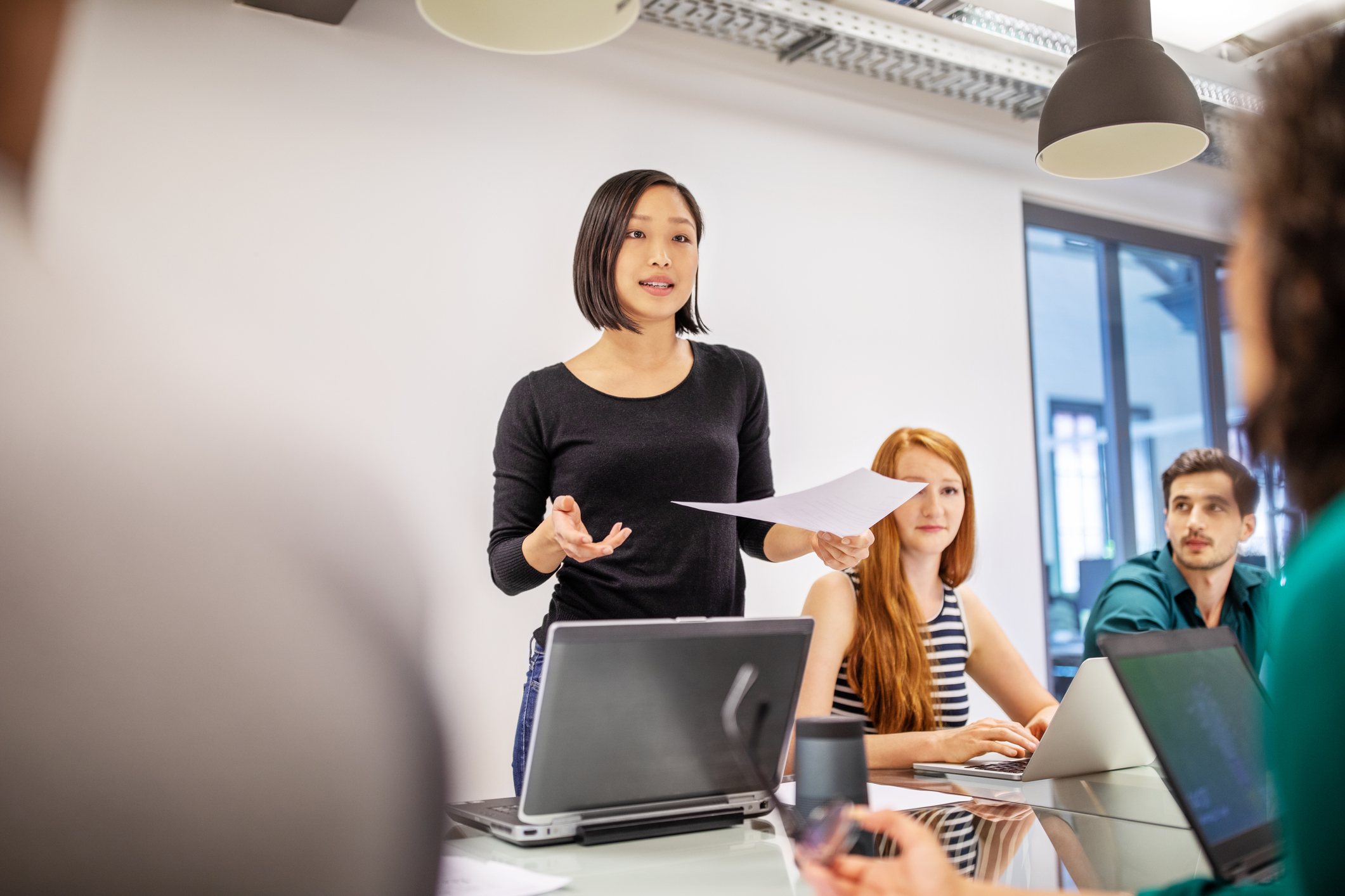 Eine deutsch-chinesische MIM Studierende mit fast schulterlangen, glatten schwarzen Haaren und schlichter dunkler Kleidung steht vor einer weißen Beamer-Wand in einem Lehrveranstaltungsraum und präsentiert etwas. Vor ihr steht ihr Notebook. Neben ihr sitzen zwei Kommilitonen ihrer Projektgruppe, eine rothaarige Studentin, die nach vorn zum Publikum schaut und ein dunkelhaariger Student, welcher zu der Präsentierenden hinaufblickt. 